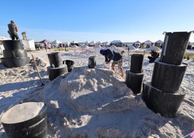 People preparing sand sculpting molds on the beach with stacked black forms and tents in the background.