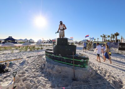 Person shaping a tiered sand boat sculpture on the beach during a sunny event.