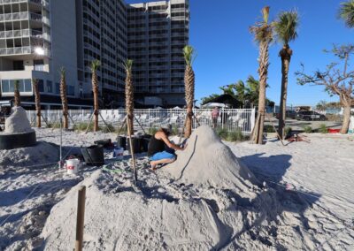 Person kneeling to carve a large sand mound with palm trees and buildings behind.
