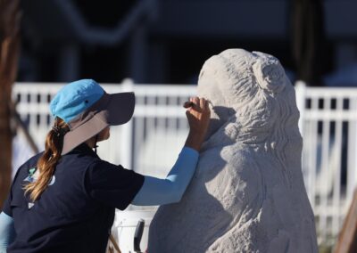 Person in a wide‑brimmed hat carving fine details into a large sand sculpture of a textured figure.