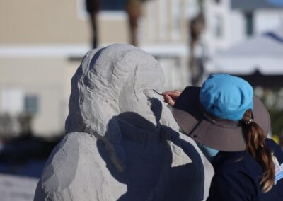 Artist in a hat carving details on a sand bust with buildings in the background.