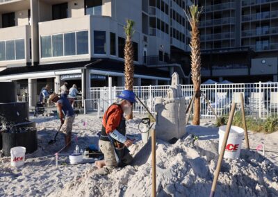 Artists shaping a sandcastle‑style sculpture near a beachfront building with tools and buckets in the sand.