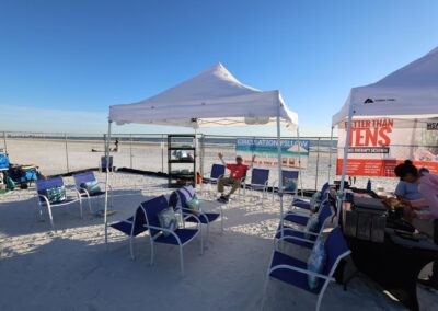Two white tents on the beach offering therapy demos, with blue chairs, banners, and staff under sunny skies.