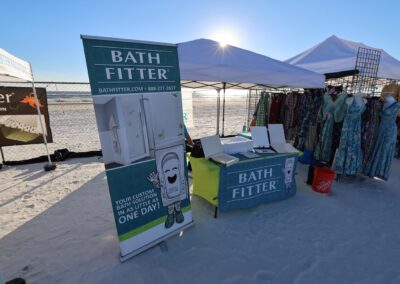 Beachside vendor tents with a Bath Fitter display table and banner beside a rack of colorful dresses.