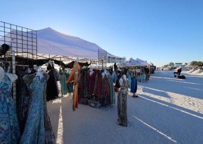 White canopy tents on the beach displaying colorful dresses on racks and mannequins in bright sunlight.