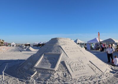 Large sand mound with carved sponsor panels at a beach festival.