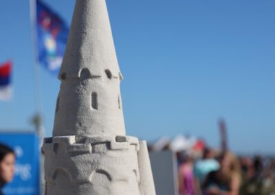 Detailed sand sculpture of a castle tower with carved brick textures and a conical roof at a beach event.