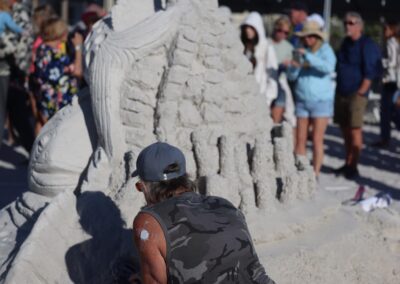 Person in a camo shirt carving an intricate sand sculpture while spectators watch and take photos.