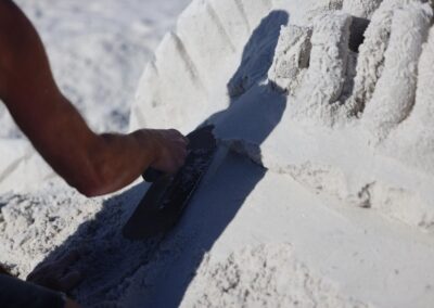 Close‑up of a hand using a flat metal tool to carve precise ridges into a sand sculpture.