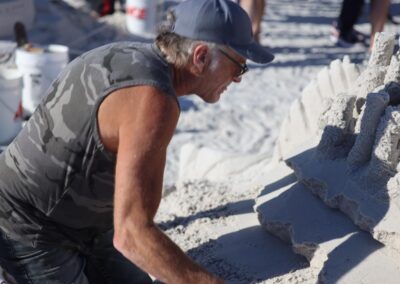 Person in a camo shirt and cap carving a layered, geometric sand sculpture on the beach.