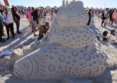 Large sand sculpture of three stacked fish with carved patterns, surrounded by spectators.