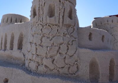 Intricate sandcastle with towers, arched windows, and a tall spire set against a clear blue sky.