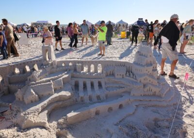 Crowds gathered around an intricate sandcastle with towers, arches, and detailed brickwork at a beach festival.