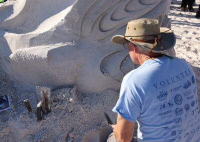 Man in a wide‑brimmed hat carving a detailed sand sculpture with tools in the sand beside them.