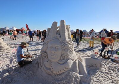 Detailed sand sculpture of a face with flowing hair and tall tower‑like forms, surrounded by artists and spectators.