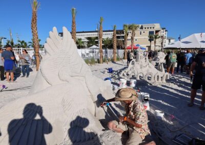 Artist shaping a large sand sculpture of birds while crowds watch at a beach festival.