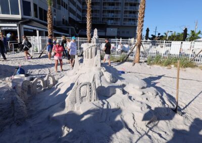 Elaborate sandcastle with towers, arches, and stairways surrounded by spectators and sculpting tools.