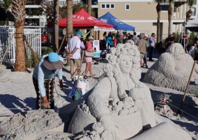 Artist shaping a sand sculpture of a curly‑haired figure with floral details as people gather nearby.