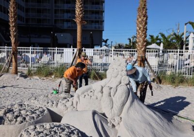 Artists working on a large sand sculpture of a seated figure with elaborate hair and clothing at a beachfront event.