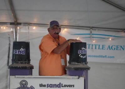 Presenter in an orange shirt giving a sand sculpting demo at a table labeled “the sand Lovers Quick Sand Speed Sculpting Show.”