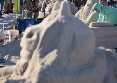 Multiple detailed sand sculptures lined up on the beach as visitors walk, watch, and take photos.