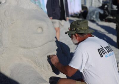 Person in a “got sand?” shirt shaping a large sand sculpture of a wide‑mouthed creature on the beach.