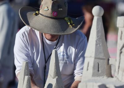 Person in a decorated hat sculpting a tall, detailed sandcastle with pointed towers at an outdoor event.