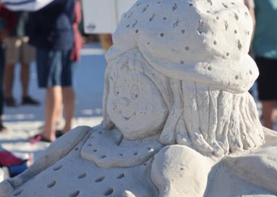Sand sculpture of a smiling character with a large strawberry hat as visitors walk among nearby tents.