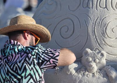 Artist in a straw hat shaping a swirling sand sculpture with the word “Sandt” carved into it.