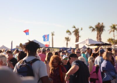 Large crowd gathered under palm trees and tents at an outdoor coastal festival on a sunny day.