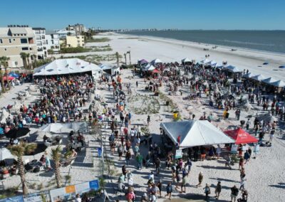 Crowds walking among white tents and sand sculptures at a large beach event under a clear sky.