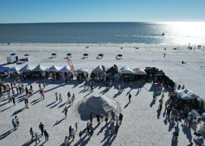 Crowds browsing vendor tents near a large sand sculpture, with blue umbrellas and ocean views in the background.