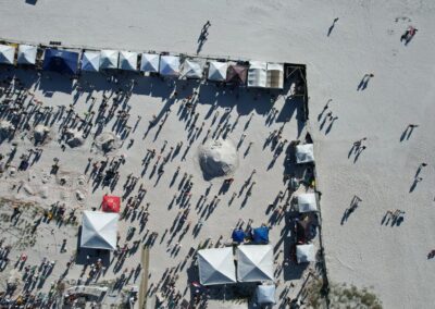 Aerial view of a busy beach event with rows of white tents, crowds, and nearby hotels and homes.
