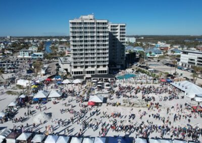 Aerial view of a busy beach festival with rows of white tents and large crowds near the shore.