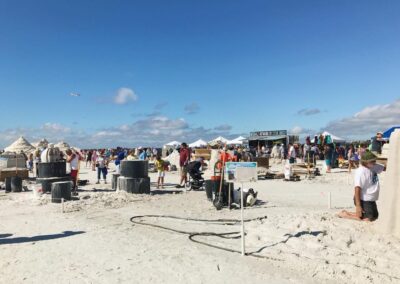 Crowds working on sand sculptures with tools, tents, and a clear sky at a beach event.