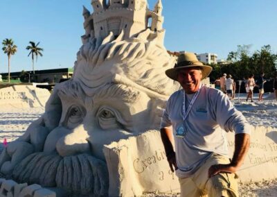 Person kneeling beside a sand sculpture of a large face topped with a detailed castle.