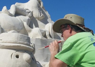 Artist in a hat brushing details into a large sand sculpture featuring multiple carved figures.