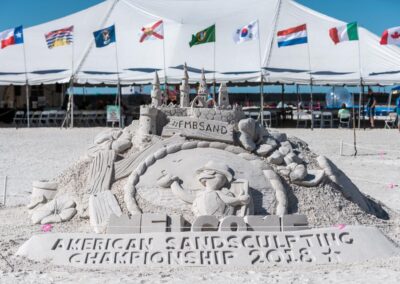 Sand sculpture with castle, seashells, and a figure in front of international flags at a sand sculpting event.