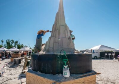 Two artists shaping a tall geometric sand tower as sunlight shines directly above its peak.