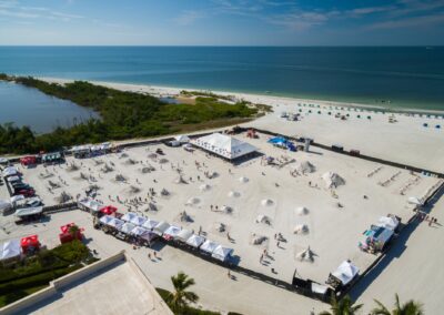 Aerial view of a fenced sand sculpting competition with tents, visitors, and ocean in the background.