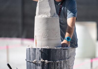 Sand sculptor shaping a tall stacked sand tower while wearing a hat, sunglasses, and gray shirt.