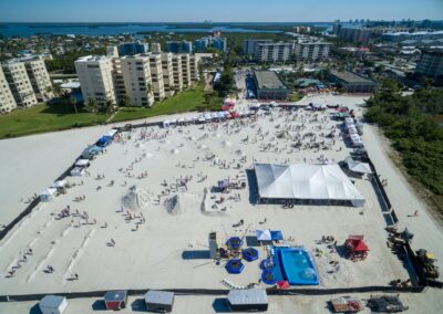 Aerial view of a beach sand sculpting festival with tents, crowds, and rows of sculptures near the shoreline.