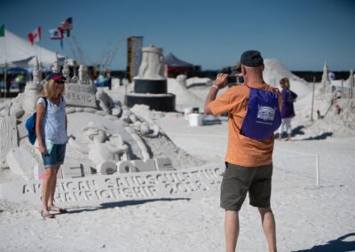 Person taking a photo of a large sand sculpture at a beach event with flags and tents in the background.