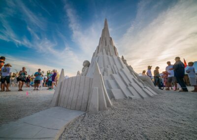 Large angular sand tower with carved figures as visitors gather around under a warm, partly cloudy sky.