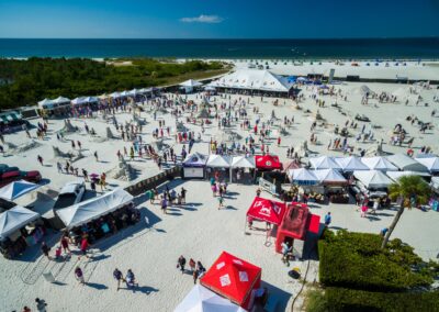Aerial view of a beach festival with tents, crowds, and rows of sand sculptures near the shoreline.