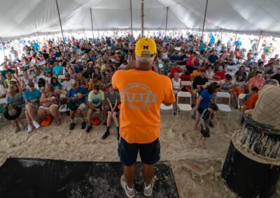 Large crowd seated under a beach tent as a staff member in an orange shirt addresses the audience.
