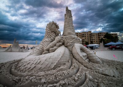 Detailed sand sculpture of a coiled dragon wrapped around a tall castle tower under dramatic skies.