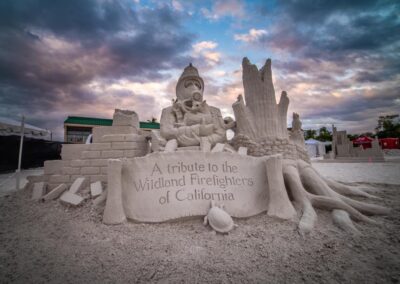 Sand sculpture of a firefighter holding a child, set against a dramatic sunset sky.