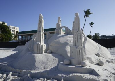 Tall geometric sand towers connected by an arched bridge, displayed outdoors with palm trees in the background.