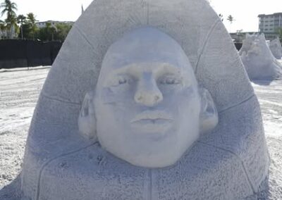 Sand sculpture of a human face emerging from a textured dome at a beach event.